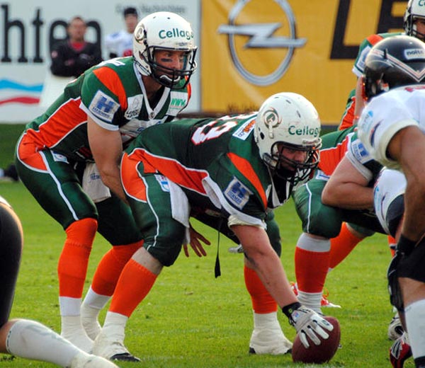 one football player is crouched behind another; the first is reaching between the legs of the second, about to receive the ball being "hiked" by him