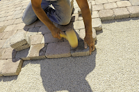 Stratenmaker aan het werk om de straat dicht te leggen, op zijn knieën en een stapel stenen naast zich.