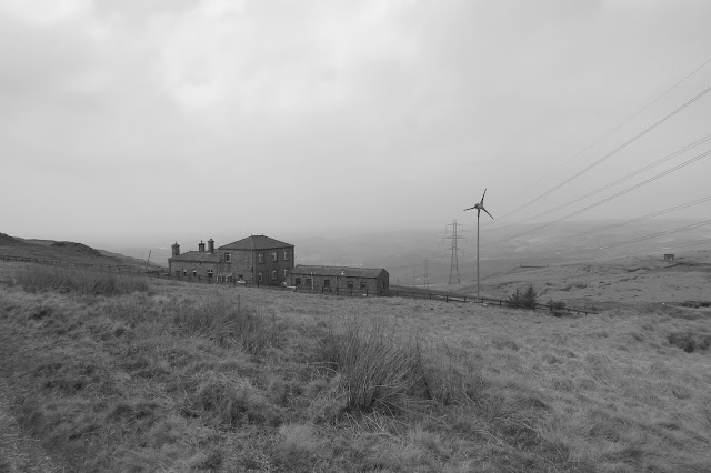 A black and white photo of a farm and a small wind generator. Wires and a line of pylons disappear into the haze of the valley.