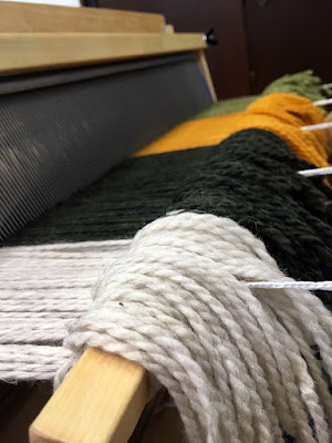 The front apron rod and reed, seen from the left at an extreme low angle. White, dark green, marigold, and light green wool yarn form broad stripes in a smooth layer emerging from the reed and tied to the apron rod. The tails pour over the apron rod to the right, and the five white cords supporting the apron rod emerge from these and continue right out of frame.