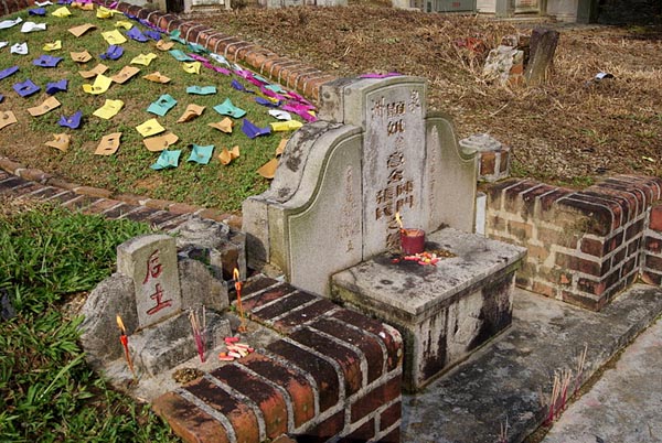 Chinese-style tombstone with offerings; mound behind is covered with colored envelopes