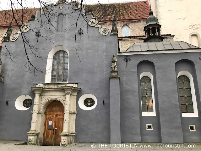 A wooden door adorned with a beautiful stone arch, and narrow and tall lattice windows on the facade of a dark grey painted facade of a church.