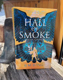 log with an axe imbedded in it and resting against its head is the book Hall of Smoke by H.M. Long with its yellow cover, blue owl, and outline of a Nordic warrior woman holding an axe