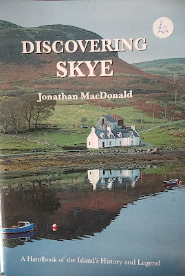 A photo of a small, glossy pamphlet entitled "Discovering Skye" by "Jonathan MacDonald", with the subtitle "A Handbook of the Island's History and Legend". The picture behind the text is of a large, gabled, slate-roofed, white-painted house sitting next to a virtually mirror-still loack with a couple of small, brightly-painted  fishing boats (dorries) and pinkish buoys on it. Behind the house stretches green moorland and a few trees, but the landscape is dominated by the sweep of dark green and russet hills which swell, round and wild and untouched except by the dottings of white sheep. A tiny road winds away, lined with old-fashioned telegraph or electricity poles. Hanging in the blue sky like an incongruous full moon is a slightly tatty, round white sticker with £2 handwritten on it in blue biro.