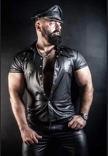 Dark bearded letter Daddy standing from the knees up wearing black leather shirt, pants and Muir cap