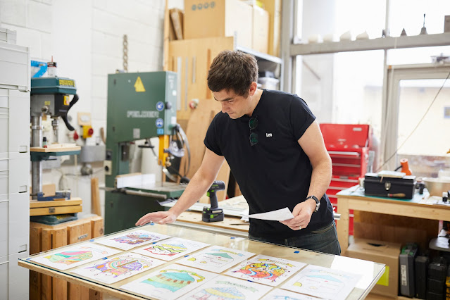 David Appleyard in his studio, reviewing colourful alternative Knottingley town crests made by local school children under a large panel of glass. There are lots of various pieces of artmaking equipment behind him.