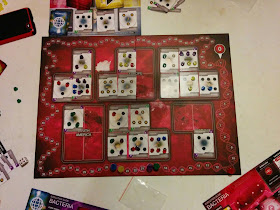 A view of the game board during a game in progress. The board consists of a scoring track around the edge, stylised to resemble a looped strand of DNA, with each 'space' made up of a section of the DNA between twists. The rest of the board is covered with twenty five spaces on which cards can be placed, overlaid on an image of the world map in red, grouped by continent: three spaces for North America, four spaces for South America, five spaces each for Europe, Asia, and Africa, and three spaces for Oceania. There are several Country cards on the board (only seven spaces are empty in total). These cards are marked with clusters of varying numbers (from three to seven) of hexagonal icons, with the icons on some of them marked with a sun symbol or a snowflake symbol. Most of the hexagonal icons are occupied by a plastic token, also hexagonal, in one of five colours: blue, green, red, yellow, or purple. Above the game board can be seen part of a play mat which contains several types of cards: a stack of yellow Event cards, a stack of red Trait cards, a stack of blue Country cards, and three additional country cards face up waiting to be chosen. Around the board can be seen some additional components (claimed or discarded Country cards, more plastic tokens, the Death Die—a standard six-sided die, white with red flecks—and portions of players' Evolution Slide play mats).