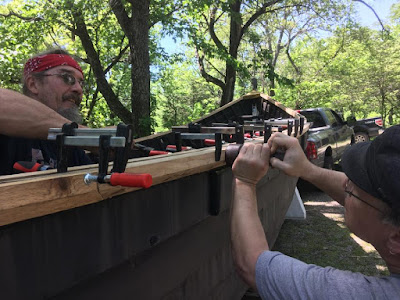 Two men working on opposite sides of a new rail on a clinker-built Viking ship, one inside and one outside the boat, with several clamps holding the rail in place.