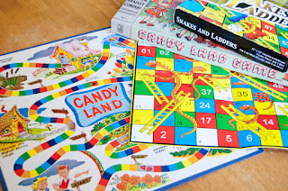 The boards for the games Candy Land and Snakes and Ladders, overlapping on a table, with the boxes for both games stacked on them. These are two of the worst examples of games with no player agency.