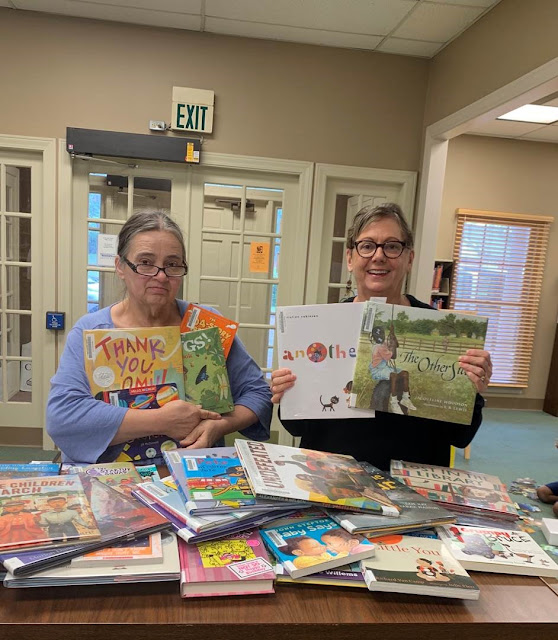 Two women each hold several picture books. Many more are piled on a table in front of them.