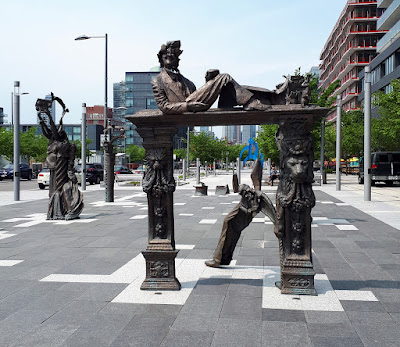 A public art piece in bronze on a broad pedestrian area made up of paver tiles. The sculpture is in several pieces, each made up of a conglomeration of disjointed parts.