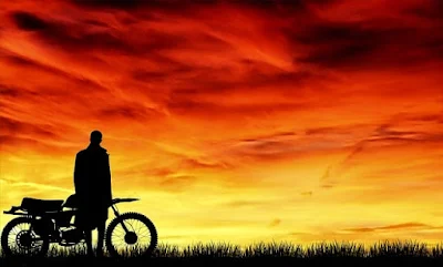 Siluet seorang pria berdiri di samping motor di bawah langit senja yang membara.