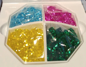 The plastic gems in their storage container in the game box. The containers are each diamond-shaped pentagons situated together so that they each form a quarter of an octagon in the box. The gems colours are yellow, green, blue, and pink.