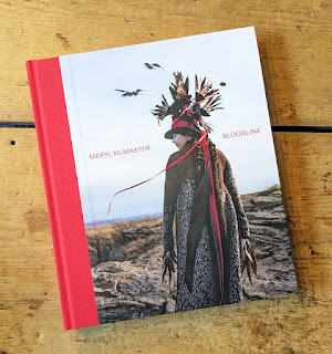 Image of cover of Meryl McMaster's Bloodline book,  showing a picture of McMaster herself, wearing a black coat with white bird foot prints over it, and a top hat with red ribbon and many feathers stuck in it,  on a rocky landscape with fields and dull blue sky behind.