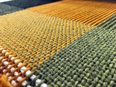 Low angle, diagonal shot of woven wool fabric, showing light green, marigold, and dark green stripes with light green weft. Three rows of white cotton yarn are just visible at lower left