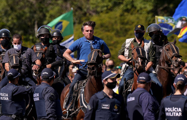 Em primeiro plano, vemos uma fileira de policiais militares uniformizados. Logo atrás deles está o presidente Jair Bolsonaro montado em um cavalo, ele está vestido com uma camisa azul e calças jeans, ao seu lado se encontram mais policiais. Ao fundo é possível notar a presença dos apoiadores do presidente vestidos de verde amarelo e balançando bandeiras do Brasil.