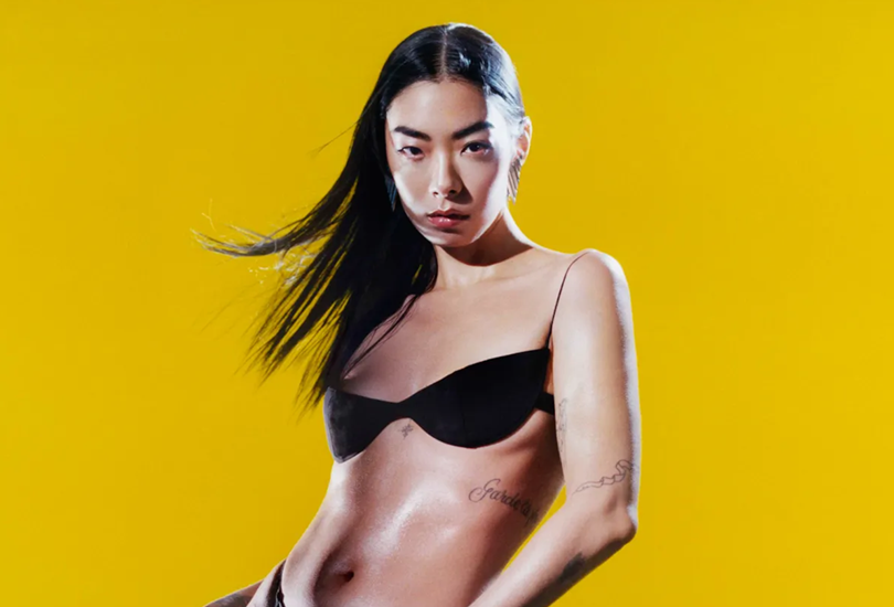 Rina Sawayama is stood in front of a yellow backdrop. Rina is wearing a black bikini top, with her hair down at her shoulders. Several of her body tattoos are clearly visible.