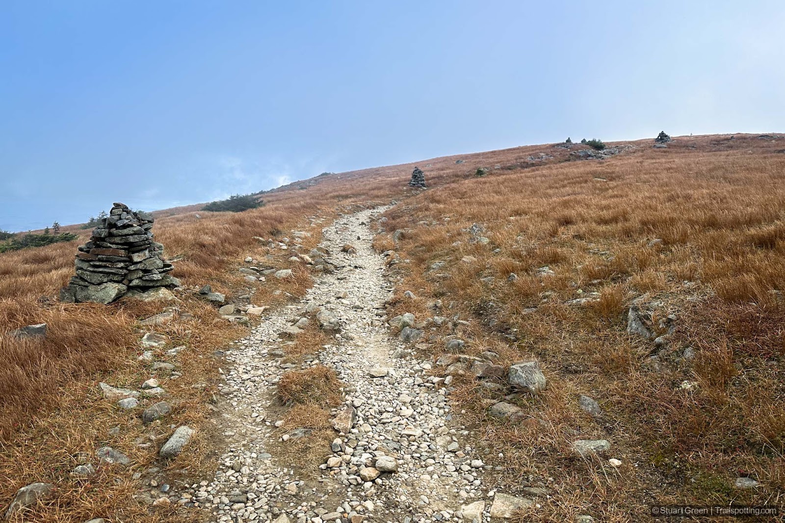 Orange grass on both sides of the trail, with sturdy rock cairns marking the route.