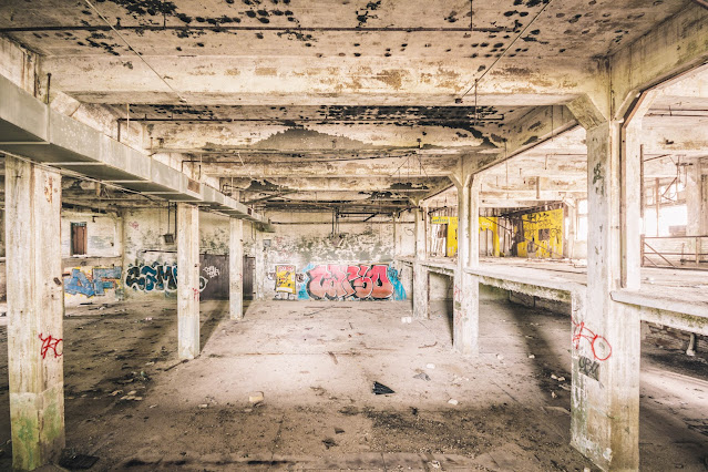 An expansive, derelict interior of a former industrial building, with rows of concrete columns and beams supporting the decaying ceiling. The area is marked by graffiti, dirt, and scattered debris, with natural light faintly illuminating the desolation and highlighting the building's forsaken state.