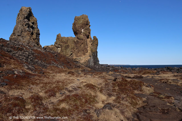 Two high basalt stacks by the ocean surrounded by rocky terrain under a bright blue cloudless sky.