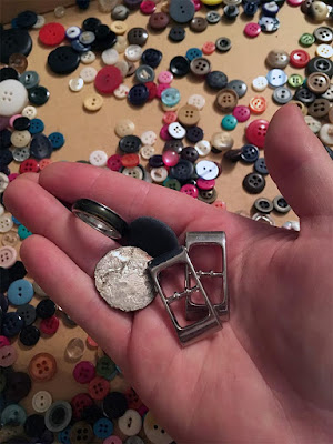 A white hand holding two rectangular silver buckles, a very battered quarter, a mood ring, and a round, flat pebble, above a variety of shirt buttons.