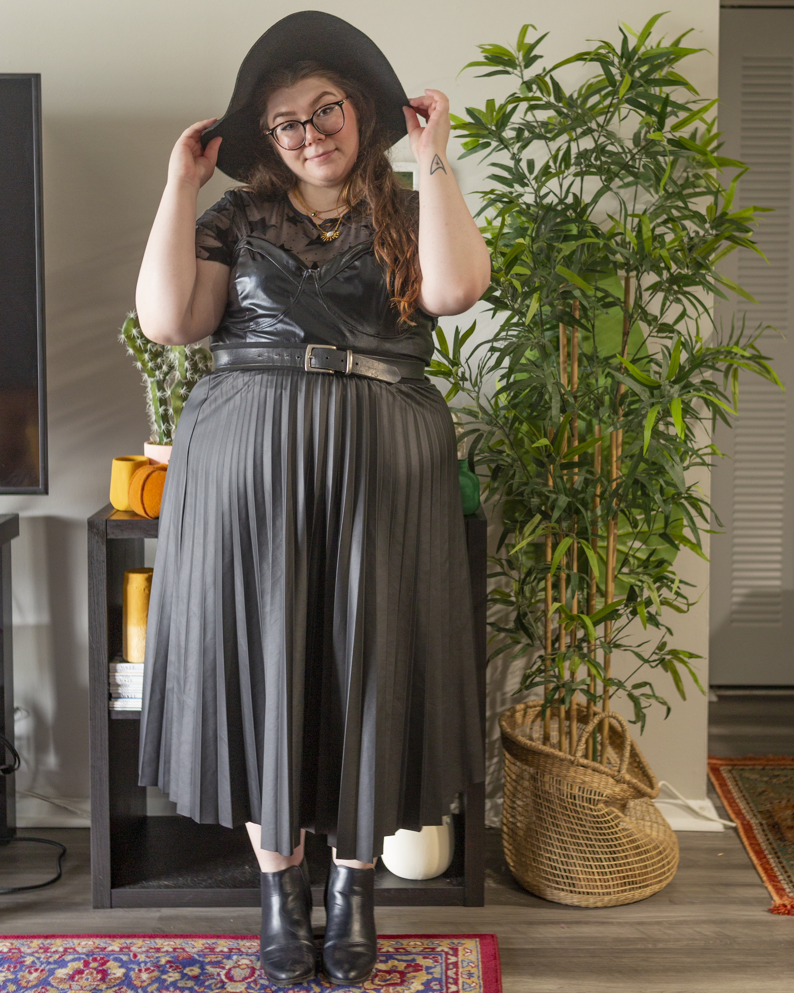 An outfit consisting of a black wide brim hat, a black sheer short sleeve tee with lettuce hems and velvet embossed starts under a black faux leather camisole crop top, a black faux leather midi skirt and black Chelsea boots.