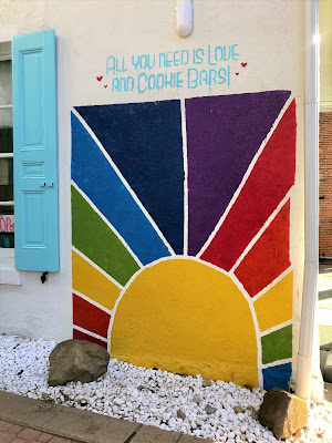 A white building with light blue shutters and a rainbow colored mural of a rising sun, with the slogan, "All you need is love, and cookie bars!"