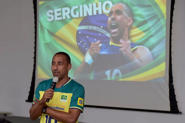 Serginho está de camisa verde e amarelo e está com microfone falando. Ele é moreno e tem cabelo ralo.