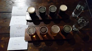 Wooden table with 2 flights of 4 small beers on wooden paddles. To the right of the beer flights are a glass of water for each. To the left of the beer flights are a list of beers on a white piece of paper with letterhead. The letterhead has the Guinness symbol and says "The Open Gate Brewery Tasting Notes"