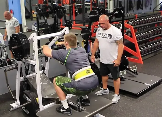 A gym instructor helps his client to do the pit shark squat