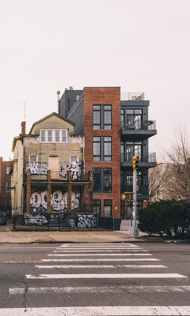 A street corner in Brooklyn showcasing the stark contrast between old and new: a graffiti-covered, dilapidated building with a weathered façade stands beside a modern, multi-story apartment with clean lines and glass balconies. A pedestrian crossing in the foreground underscores the crossroads of urban change and gentrification.