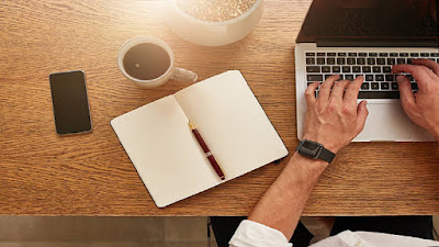 Brown top small business marketing work station with a computer, notepad and pen, smartphone and a white mug of beverage.