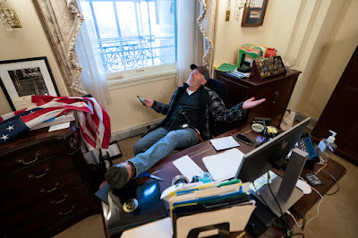 Richard Barnett kicks up his feet at Nancy Pelosi's desk, leans back, and holds his smartphone.