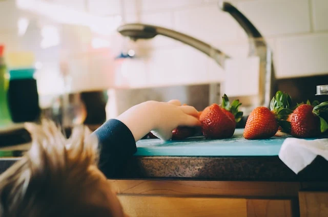A child reaching up to a kitchen surface with strawberries on a chopping board