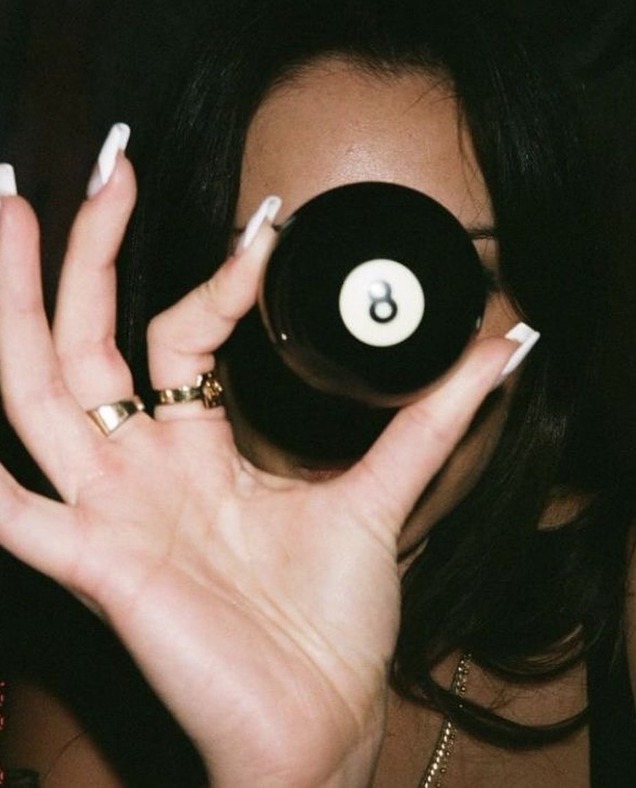 Woman with dark brown hair and 2000s Y2K French tip manicure wearing gold rings and holding an 8 ball