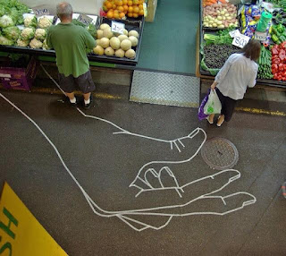 A foto vista de cima para baixo mostra o corredor de um mercado; ao centro, uma passagem estreita com uma rampa acessível. À esquerda, um homem em frente a um balcão expositor repleto de alfaces, couves-flores, melões e laranjas e à direita, uma mulher, segurando com a mão esquerda, duas sacolas plásticas e a outra próxima a outro balcão, com pepinos, jabuticabas, maços de salsões, pimentões verdes e vermelhos, berinjelas, batatas e cenouras. No chão, um enorme desenho em giz branco, de parte de um antebraço e mão direita. O antebraço parte da base da banca esquerda, por onde o pé esquerdo do homem pisa sobre o contorno interno, próximo ao punho. A mão está ao centro, os dedos polegar e indicador posicionam-se atrás da mulher, onde há uma tampa fluvial redonda, como se fossem girar a tampa. 