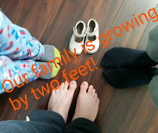 Picture of three sets of feet and an empty pair of shoes with orange text across saying, "our family is growing by two feet" by OneQuarterMama.ca