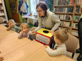 Pani bibliotekarka stoi przy grupie dzieci, trzyma w ręku kolorowe kartonowe pudełko z napisem: Magiczne pudełko.