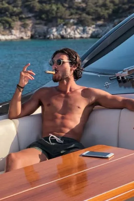 Shirtless muscular tan sunglasses wearing cigar smoking male sex object on a boat deck