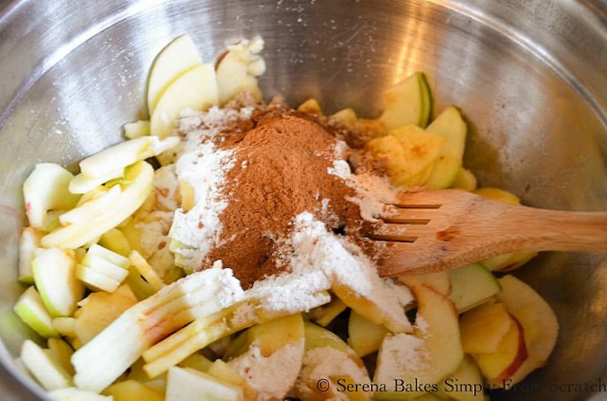 Sliced Granny Smith Apples, Braeburn Apples, Brown Sugar, Flour, Cinnamon, Vanilla and Lemon Juice in a stainless steel bowl with a wooden spoon resting against the edge.