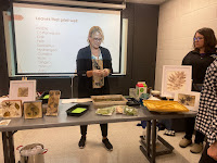 a blonde woman stands behind a table with her botanical artwork. She is explaining her process.