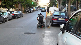 ανάπηρος με αμαξίδιο και η συνοδός του ανηφορίζουν ένα δρόμο κινούμενοι στη λωρίδα ανόδου. Παρκαρισμένα αυτοκίνητα και από τις δύο πλευρές του δρόμου δε αφήνουν να δούμε το (απροσπέλαστο) πεζοδρόμιο.