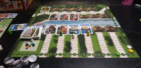 The game board, with a game in progress. The board has two sections: on the top, two rows of huts serve as places for cards. The top row has only one card, with a yellow meeple on it. The bottom row has three cards, with no meeples. The bottom half of the board has a row of huts, four of which have cards on them. In front of each hut is a path divided into spaces leading up to the hut. The path leading to the first hut is empty. There is a single green meeple on the path to the second hut. The third hut has both a green and a white meeple on the path. The fourth hut has a yellow meeple on its path. The cards have artwork representing Viking warriors, crafters, traders, and sailing ships. The cards representing ships have one to three wooden cubes of different colours on them. In the corner of the board is a space for the deck of cards, and another space holding several additional coloured wooden cubes. Along the edge of the board is a scoring track, with a green, white, and yellow disc at various points along it. On the table near the board are a stack of tokens representing coins and a player's board with some unused meeples and a variety of cards around it.