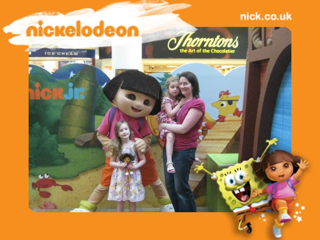 Theme park picture with text Nickelodeon and mum Steph standing holding Sasha, with a lifesize Dora holding shoulders of Tamsin