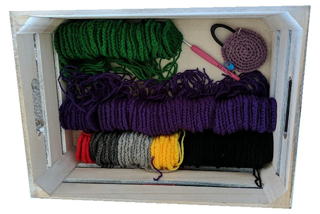 Wooden box with small crochet squares lined up in columns by colour: purple, green, red, yellow, black and grey.