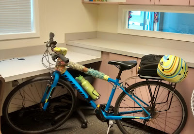 My blue Specialized Sirrus Sport bicycle with yellow and multi-striped bicycle helmet clipped to the trunkbag, parked in front of cupboards and counter-tops in an office-type setting