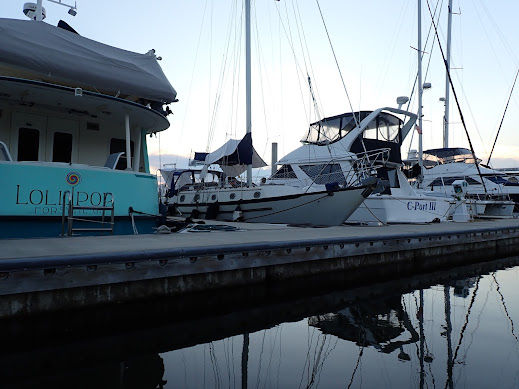 A relatively small white sailboat, Fulmar, is tied up in a slip between two large boats.