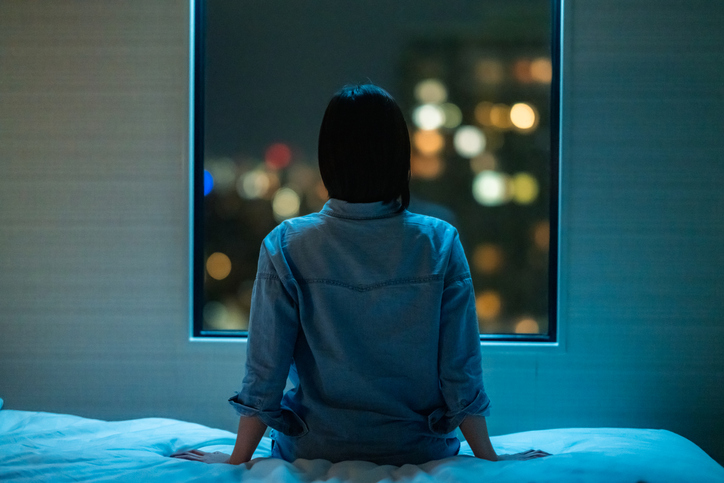 Woman sitting on a bed, back to us, facing a window showing a night time scenery
