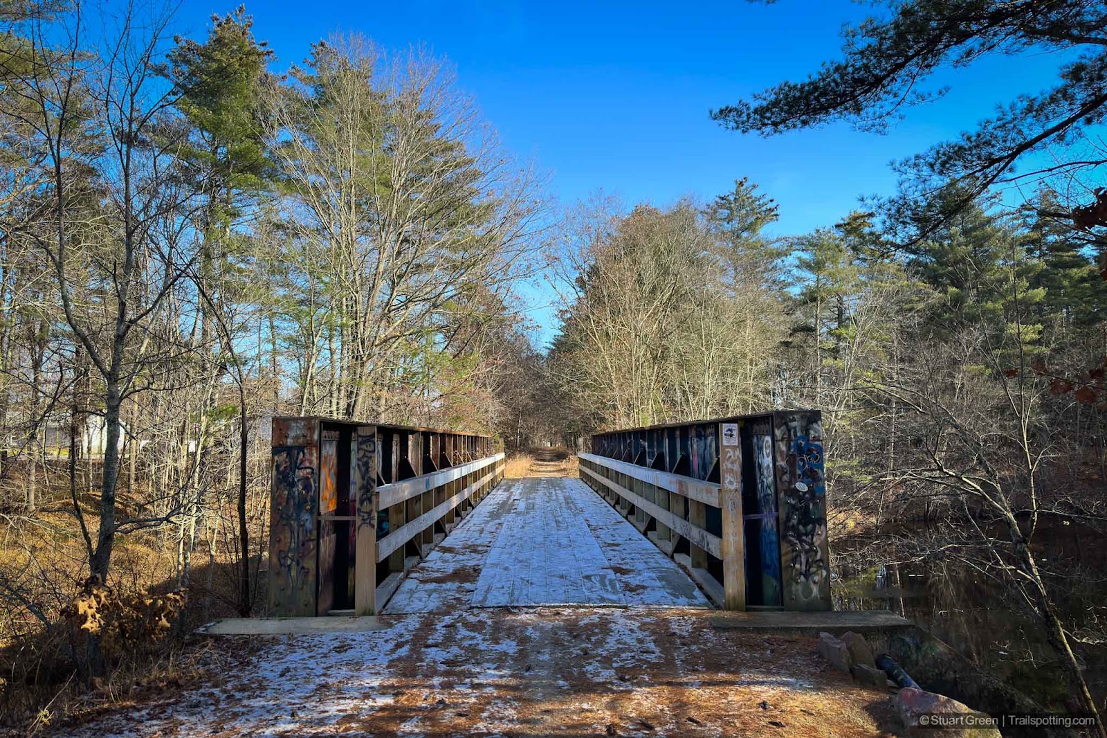 View of the bridge from above, looking straight down the trail. Some frost on the bridge deck. Graffiti on the steel girders.