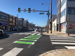 The intersection of 5th Street and Washington Avenue in South Philly.  A traffic light hangs overhead and there are green stripes through the crosswalk, indicating a bike lane.